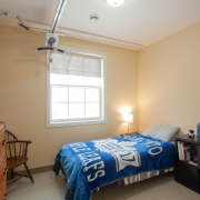 Well lit bedroom with a large window and single bed with a blue Maple Leafs bedspread. There is a wooden end table with a lamp, a dressor and brown sitting chair nestled in the corner.