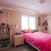 Client's bedroom has bookshelf filled with various books and stuffed animals on top; a beige night stand with a lamp, personal products and radio on top; a single bed underneath a window with a bright pink duvet with pillows and stuffed animals on top of the covers; double closet doors on right side of the room; a hoyer lift and belt attached to the ceiling for transfer support.