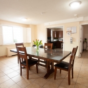 Beige dining area with rectangle brown table and four chairs. Green plant sits in the middle of the table. Well lit room is sparcely furnished and leads to a kitchen at the back.