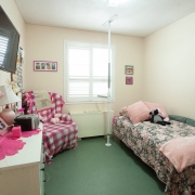 This client's bedroom has a White dressor with pink accents including lamp, radio and picture frames on the left side of the room; TV mounted above; Pink plaid lazy boy chair in top corner; single bed with pink pillows and floral duvet along the right side of the room with assist lift pole.