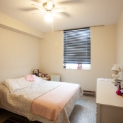 Client's bedroom has a double bed on the left hand side covered with a white floral bedspread and pink blanket at the end of the bed. A light wood dressor is across from the bed on the right hand side and is decorated with a lamp and personal items. A window with a blind is at the back of the room and a bed side table with small items is situated in the back corner on the left hand side. There is a white ceiling light with a fan.