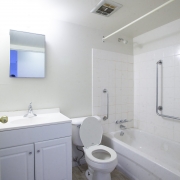 All white bathroom has a white vanity with 2 doors to the left and a mirror hanging above. A white toilet is in the middle and a white bathtub shower unit surronded by white tiles is situated to the right. The bathtub is equipped with 2 safety bar handles, a white curtain rod is attached to the wall, and a fan/vent is installed on the ceiling.