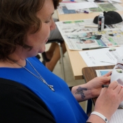 Female client sitting at a table, holding a white mask that says 'STAY STRONG' on the forehead; she is designing the mask as part of a creative expression workshop with the intention of reflecting on her personal brain injury journey.