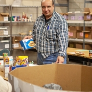 Male client is standing in the middle of a large wearhouse in front of a large open cardboard box. There are several open shelving units along the back that are housed with food and other boxes. The client is holding a box of food in his right hand and looking at the camera. He is organizing food donations at food bank during volunteer placement.