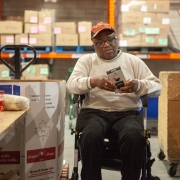 Male client in a wheel chair is sitting in the middle of a wearhouse looking at a brochure. There are several large boxes around him, and orange metal shelving units are lined up against the back. Small labelled boxes in taped together groupings are housed on the shelves behind him.