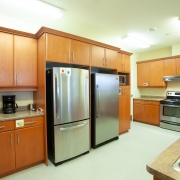 Large kitchen with wall of brown wood upper and lower cabinetry, beige walls and light flooring. 2 stainless steel fridges side by side in the middle. Beige countertops with a coffee pot on the counter to the left of the fridges and a microwave insert in the cabinetry to the right of the fridges. Another wall of brown wood cabinetry with upper and lower storage along the back wall with a stainles steel oven, black cook top and fan installed above.