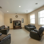 Living room has 2 dark brown lazy boy chairs and 2 brown couches that are set up to watch the television set up on a dark brown television unit. The walls are painted beige and there are 2 large bright windows letting in a lot of natural light. Above the tv is a clock and 2 wall hangings are on either side of the tv stand.