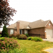 Brown brick bungalow with double car garage; accessible ramp on the left side leading up to the front door; well-maintained front lawn with trees and bushes