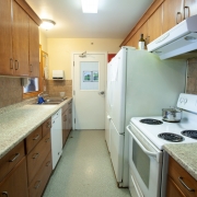 Long narrow kitchen with light brown cabinets, above and below counter top on the left side; entrance door at the back; white fridge, stove and additional counter space and cabinets on the right hand side.