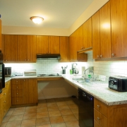 Brown upper and lower cabinetry in a narrow u-shaped kitchen. Counter tops are light coloured marbled and the back splash is white tiles. Lower cabinet units are minimized under the sink and cooktop to make this a wheelchair friendly work space. Black dishwasher is installed on the right hand side and flooring is beige tiles.