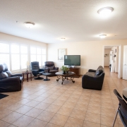 Large living room area is well lit with four ceiling lights and has a wall of windows on the left hand side letting in natural light. Walls are beige and the flooring is tiled brown. Three black lazy boy chairs, are set up along the left with an end table and foot stool beside the middle chair. A black couch is situated on the right and a round coffee table sits in the middle between the couch and chairs. The coffe table has a green plant placed in the middle. A television sits on a brown wooden tv stand at the back of the room.