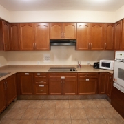 U-shaped kitched has medium brown upper and lower cabinetry. A black cook top with a fan above is set at the back in the middle of the room. A white wall stove is on the right hand side, and a white microwave sits on the counter at the back on the right hand side. Counter tops are light coloured and flooring is tiled beige.
