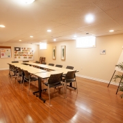 Bright basement meeting room with hardwood floors, beige walls and 2 windows. Large 12-14 person meeting tables with chairs set up in the middle of the room with a free standing easel and notice boards on the wall.