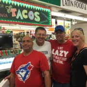 3 male clients and 1 female staff standing together and smiling in front of a Taco place at a local market during lunch.