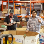 2 male clients are standing in front of large orange metal shelving units in the middle of a wearhouse. They are standing behind an empty, waist high cardboard box. They are organizing food donations at a food bank during volunteer placement.