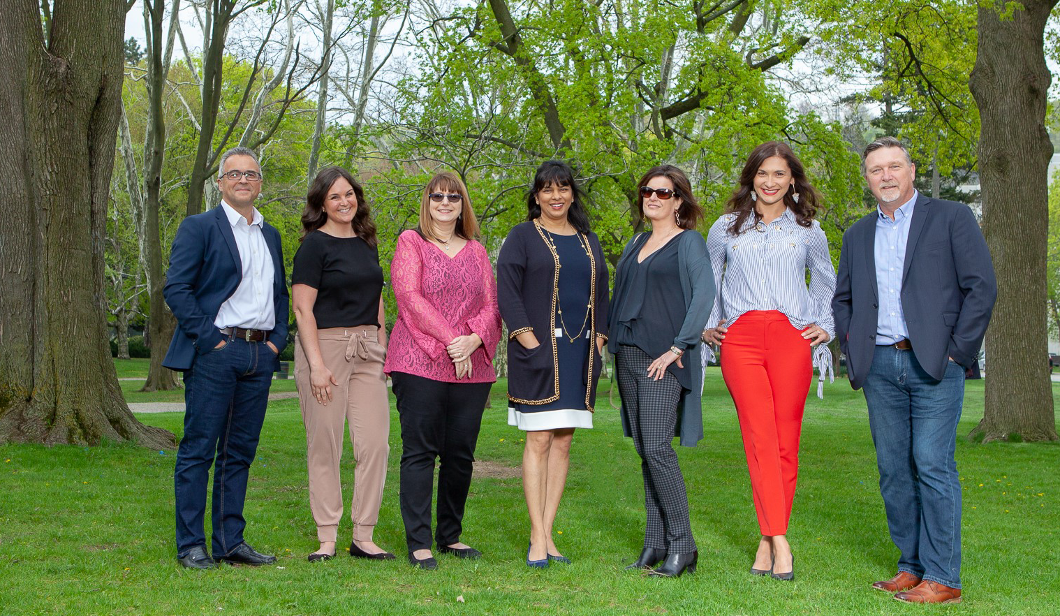 Leadership team from left to right: Vlad Bartchouk, Mary Jackson, Nadina Salciccioli, Mila Ray-Daniels, Candy Sarraf, Joanna Pajak, Steve Dawn. Staff members are standing in a row on lush green grass in a park like setting, surrounded by many large trees.